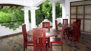 A covered outdoor dining area with wooden tables and chairs, surrounded by white columns and a view of lush greenery. - The Cave Kithulgala (Kithulgala)