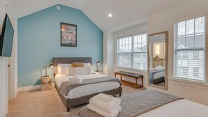 A bedroom with a large bed, bedside lamps, a bench, a mirror, and a window with blinds.