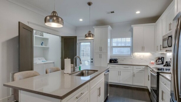 A modern kitchen with white cabinets, stainless steel appliances, a central island with a sink, pendant lighting, and a window providing natural light.