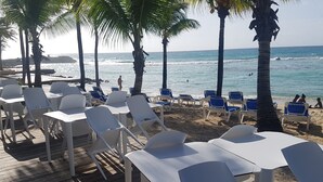 Une terrasse au bord de la plage, avec des chaises et des tables en plastique blanc, des palmiers et l’océan en arrière-plan.