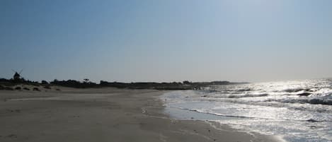 A beach with a windmill in the distance, calm water, and a clear sky.