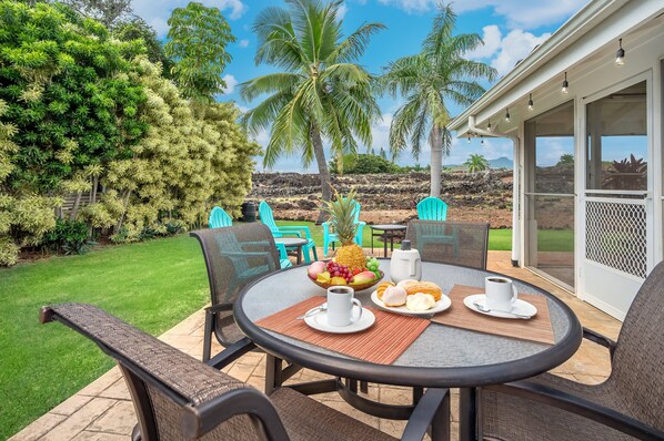 A round table with chairs and a fruit bowl, a coffee cup, and a plate of pastries on a patio with a view of a garden and a building.