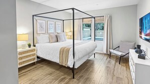 A modern bedroom with a four-poster bed, a bedside table with a lamp, a chair, and a desk with a vase.