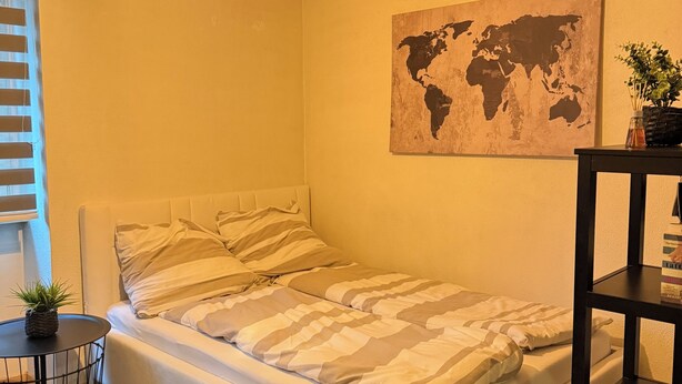 Una cama individual con una colcha a rayas, un mapa mundial en la pared, una bombilla colgante y una pequeña mesa con una planta y libros.