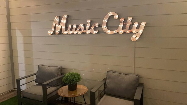 A lounge area with two grey cushioned chairs, a small round table with a potted plant, and a wall-mounted sign that reads "Music City".