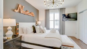 A modern bedroom with a large bed, bedside lamps, a chandelier, a TV, and a decorative wall piece.