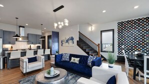A modern living room with a blue sofa, white armchairs, and a coffee table. There is a kitchen area with grey cabinets and a dining area with a black and white patterned wall.