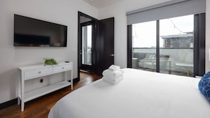 A modern hotel room with a large bed, a flat-screen TV mounted on the wall, a white console table with a plant, and a balcony with a glass door.