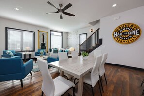 A modern living room with a dining area, featuring a large wooden table, white chairs, and blue sofas. A staircase with a black railing leads to another level. - Two Townhomes- Rooftop Lounge- Hot Tub- City Views (Nashville)