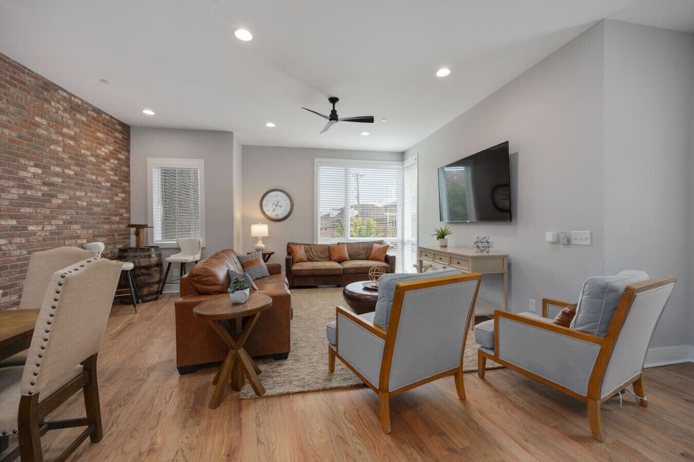 A spacious living room with a brick wall, a ceiling fan, a large window with blinds, a sofa, a coffee table, and a TV mounted on the wall.