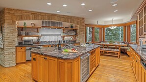 A modern kitchen with a stone accent wall, wooden cabinetry, and a large island with a built-in sink. - Large Ski-in/out Retreat + Lap Pool | Founders Lodge (Beaver Creek)