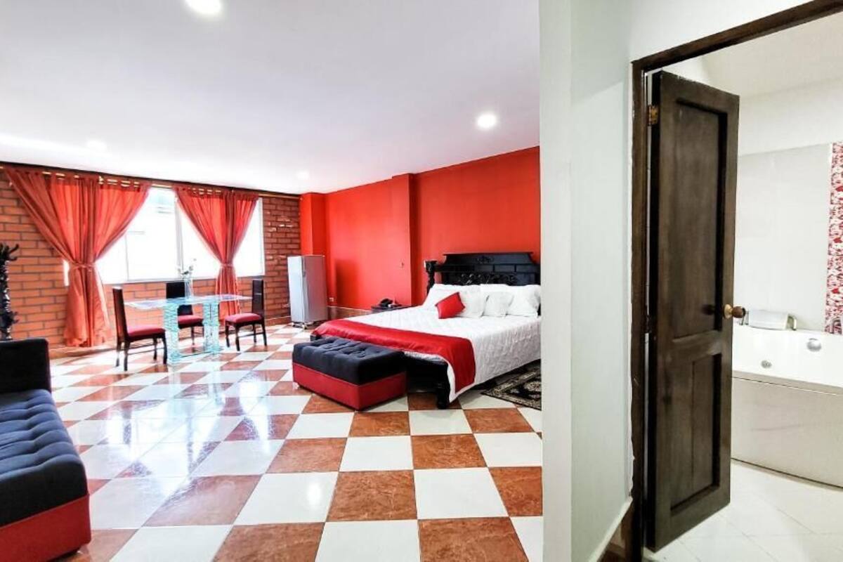 Ein Hotelzimmer mit Schachbrettfußboden, einem Bett mit roß-weißer Bettwäsche, einer roten Couch, einem Schreibtisch mit Stuhl und einem großen Fenster mit roten Vorhängen.