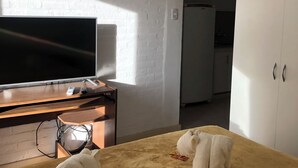 Una habitación con una televisión de pantalla plana grande sobre un escritorio de madera, un armario blanco y una cama con una manta amarilla.