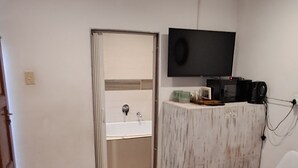 A bedroom with a bed, a TV mounted on the wall, a bathroom with a sink and mirror, and a door leading to another room.