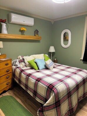 A bedroom with a plaid bedspread, a wooden dresser, a green rug, a wall-mounted air conditioner, a shelf with flowers, and a mirror.