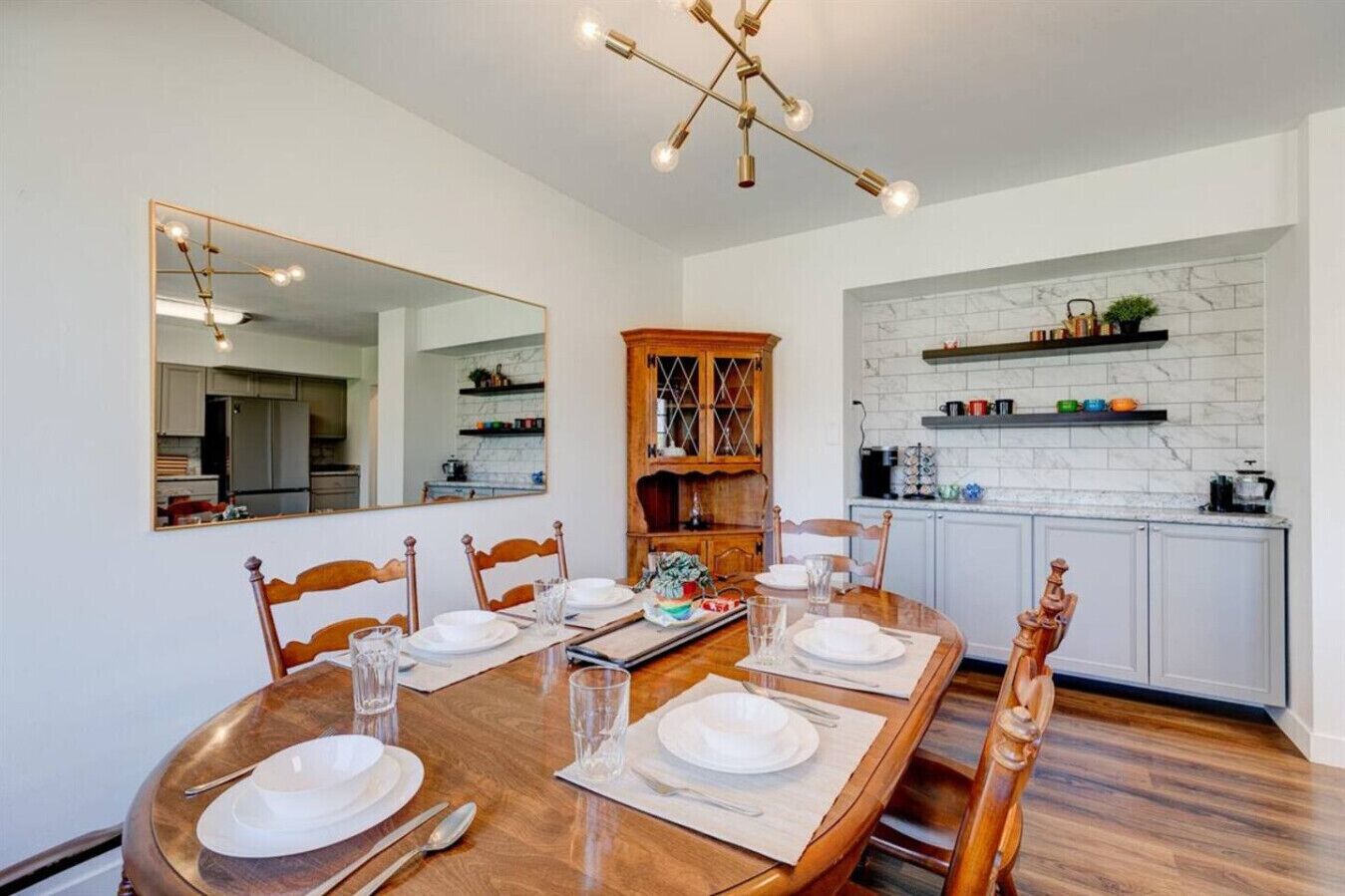 Un comedor con mesa de madera preparada para cuatro personas, un espejo grande y una cocina con salpicadero de mármol blanco y armarios de madera.