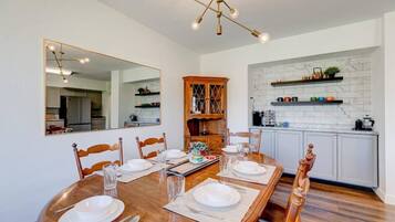 Un comedor con mesa de madera preparada para cuatro personas, un espejo grande y una cocina con salpicadero de mármol blanco y armarios de madera.