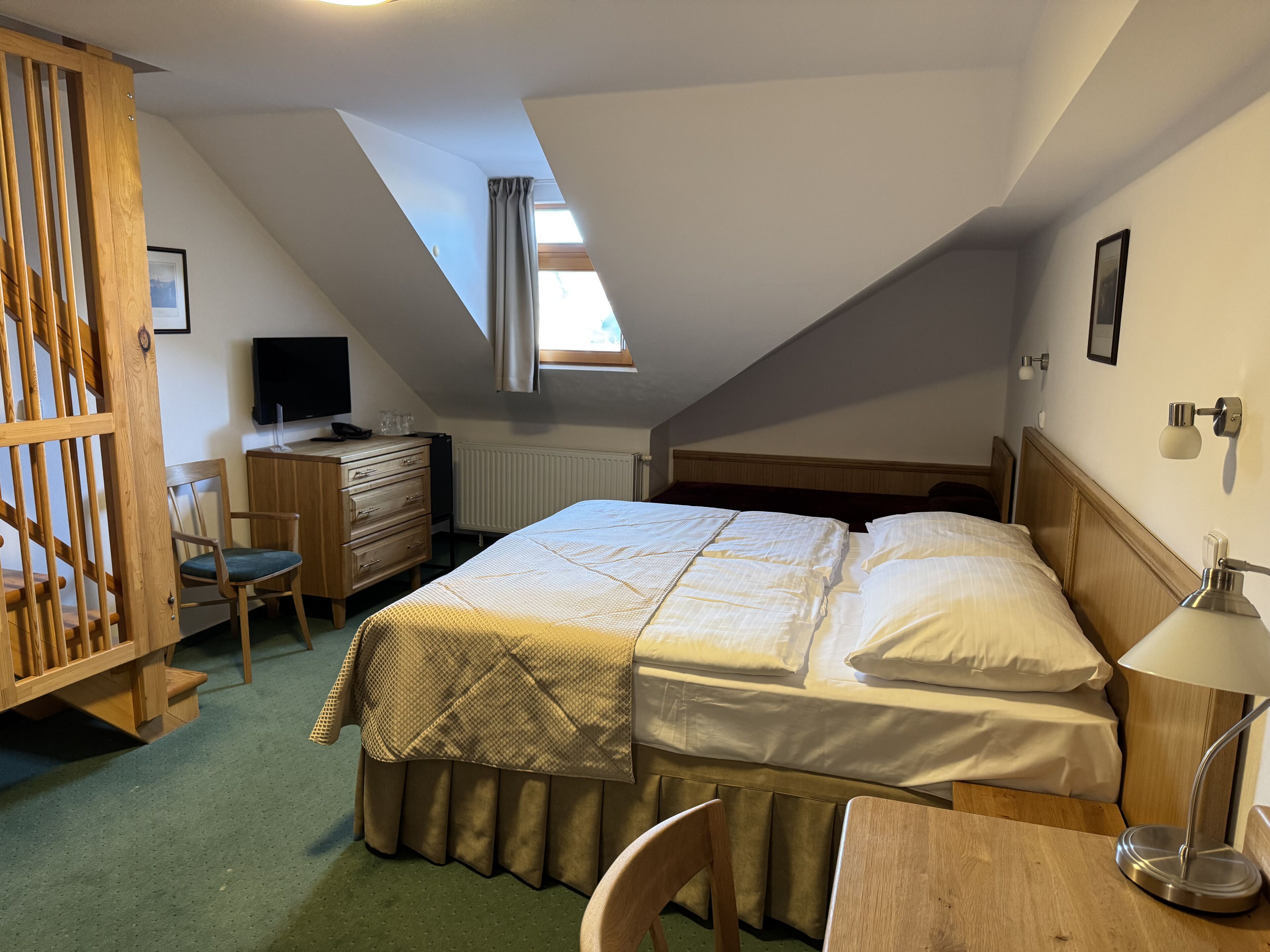 A bedroom with a bed, a desk, a chair, a dresser, a television, and a window.