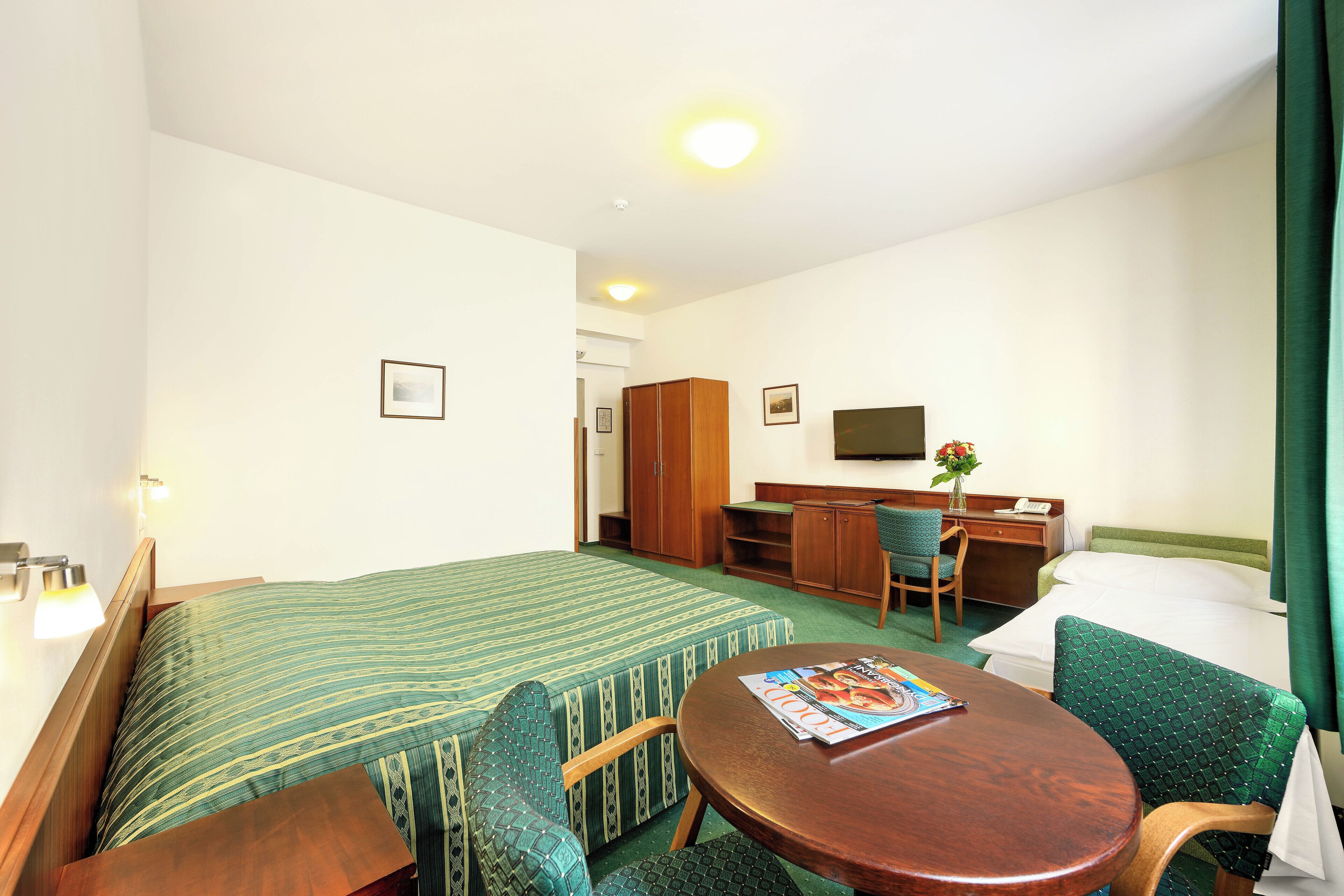 A hotel room with a bed, two chairs, a small table, a desk, a television, and a green curtain.
