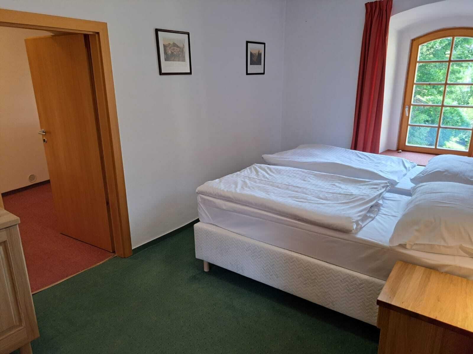 Ein Zimmer mit zwei Betten, einer Holztür, einem Fenster mit Aussicht und zwei gerahmten Bildern an der Wand.