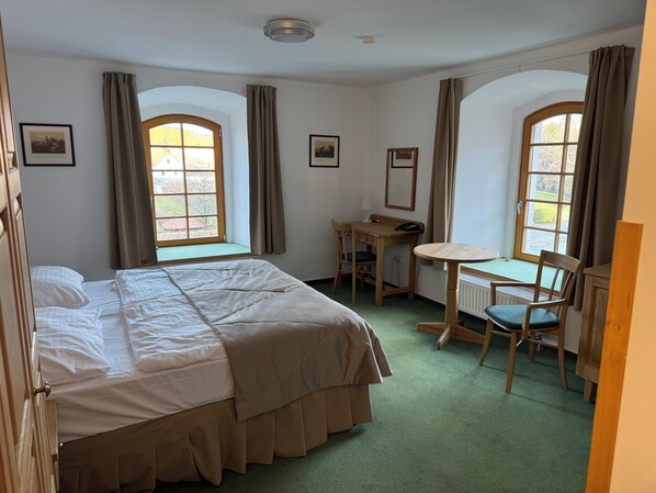 Habitación de hotel con cama, escritorio, silla y dos ventanas con cortinas.