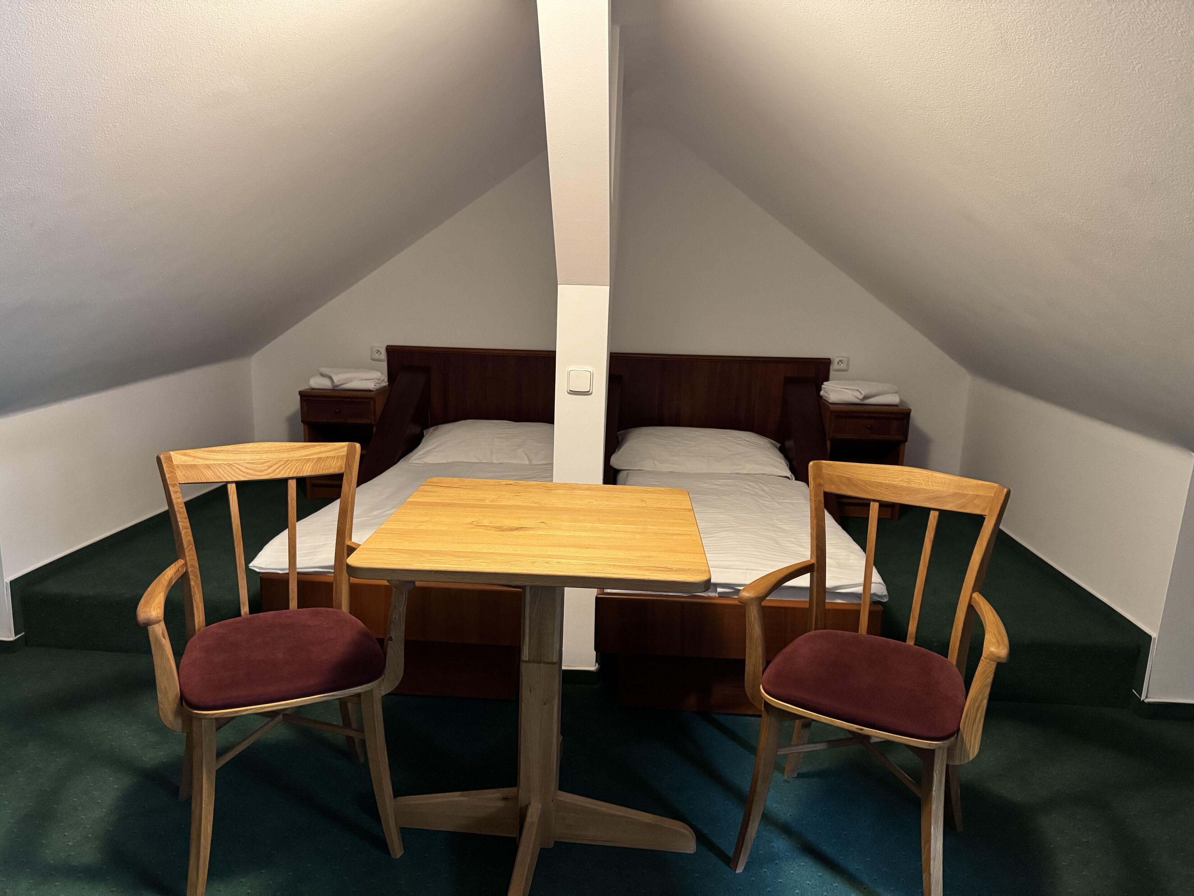 A small, triangular room with a bed, two wooden chairs with red cushions, a wooden table, and a nightstand with a lamp.