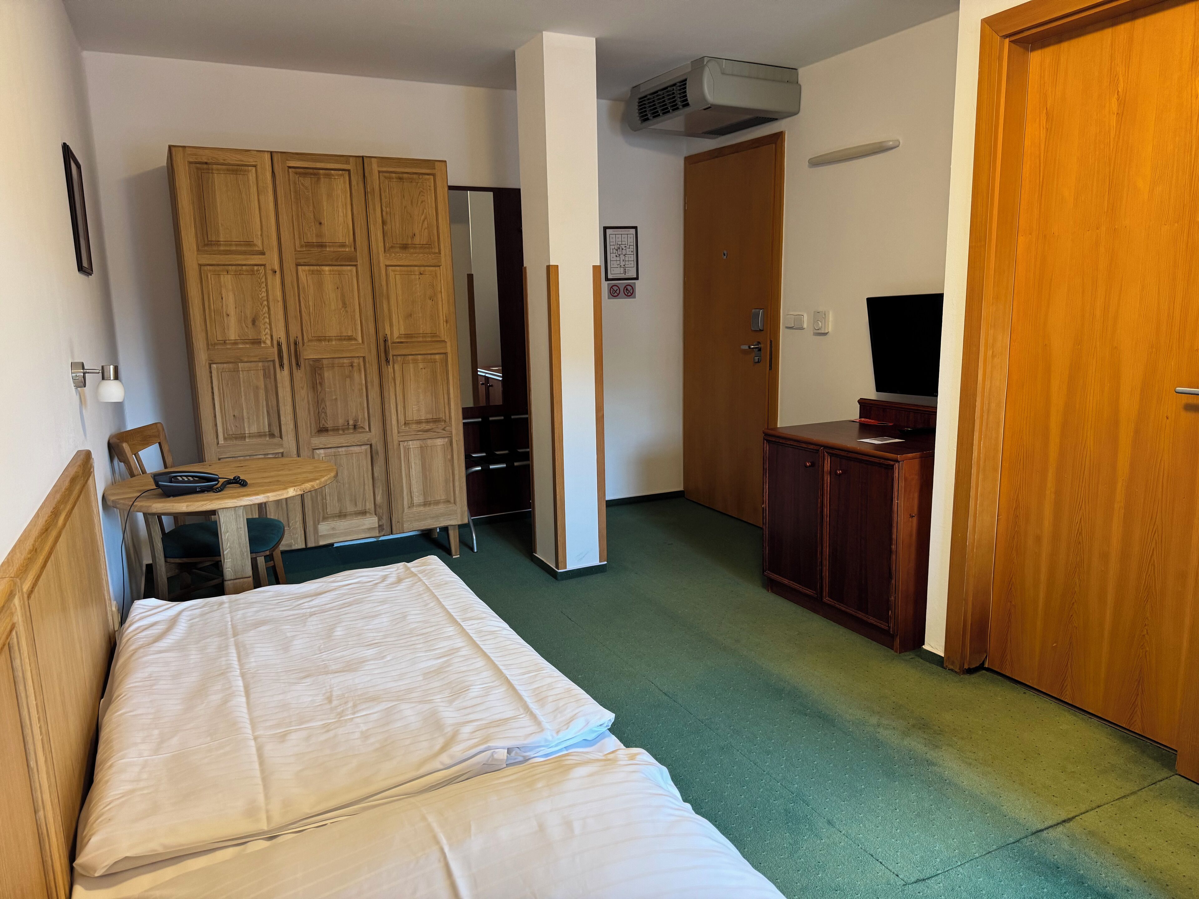 A hotel room with a bed, a wooden wardrobe, a desk with a chair, a television, and an air conditioning unit.