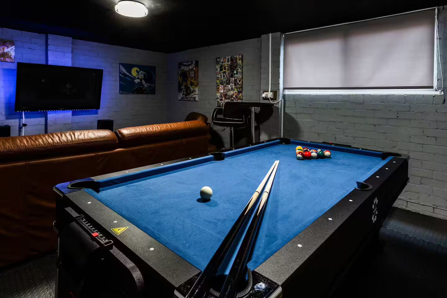 A pool table with a white cue ball and several colored balls, a brown leather couch, and a television mounted on the wall.