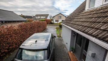 Une allée menant à une résidence, avec une voiture garée, une haie et des maisons en arrière-plan.