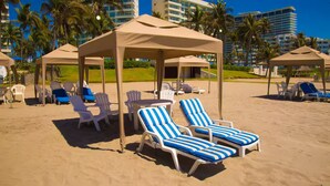 Una playa con sillas de descanso y sombrillas, palmeras y un hotel al fondo.