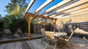 An outdoor wooden deck with a pergola, a table with chairs, and a view of a stone building in the background.