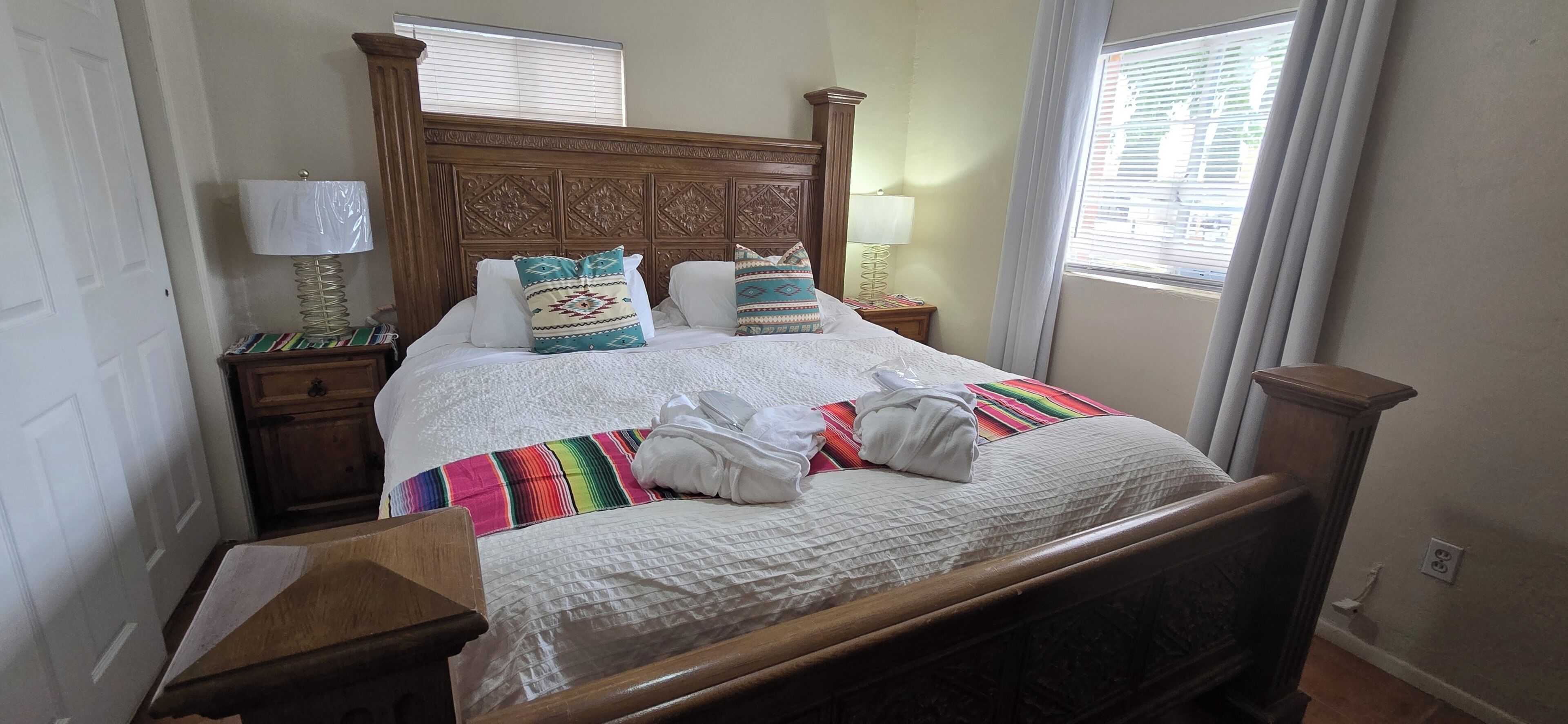 A neatly made bed with a patterned bedspread, two pillows, and a folded towel. There are two bedside tables with lamps, a wooden headboard, and a window with blinds.