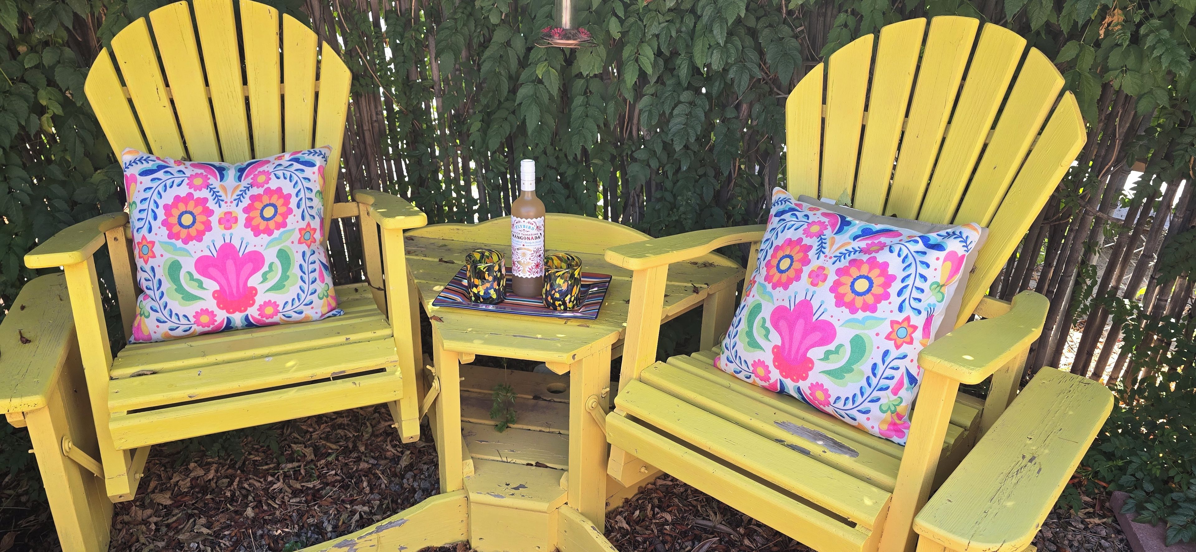 Two yellow Adirondack chairs with floral cushions, a small table with a bottle and glasses, and a decorative hanging in a garden setting.