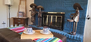 A room with a blue brick fireplace, a wooden side table with books, a colorful striped blanket, and two ceramic cups on a tray.