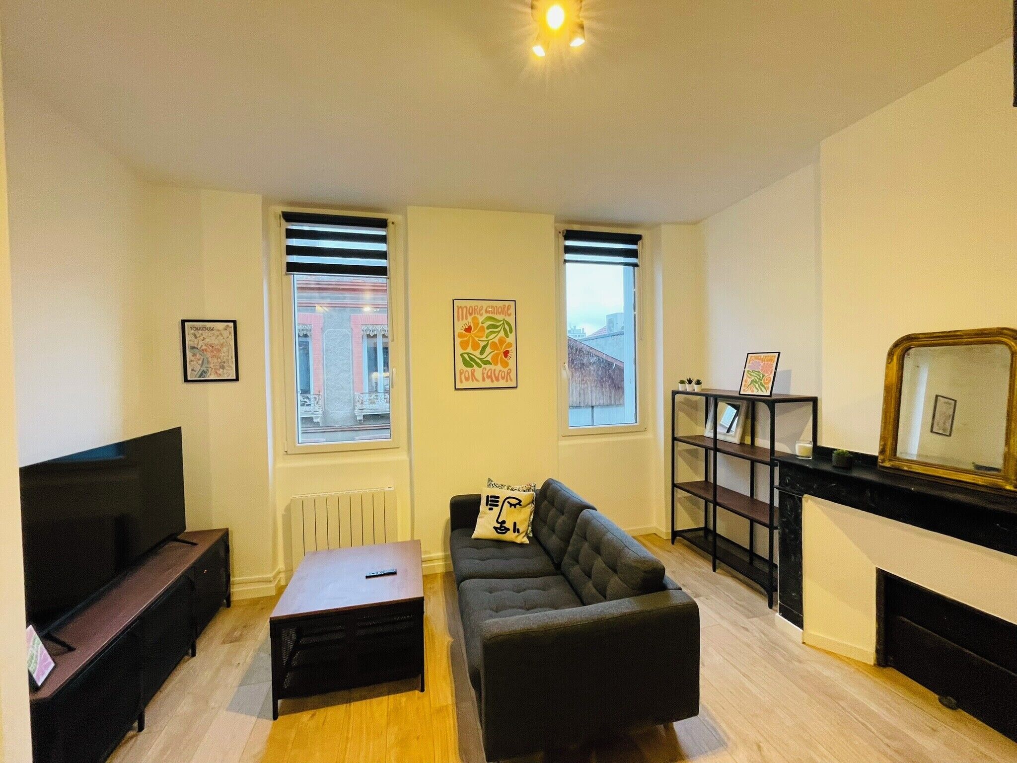 Una sala de estar con sofá, una mesa de centro, televisor y chimenea. Hay dos ventanas con persianas, y una estantería con fotografías enmarcadas.