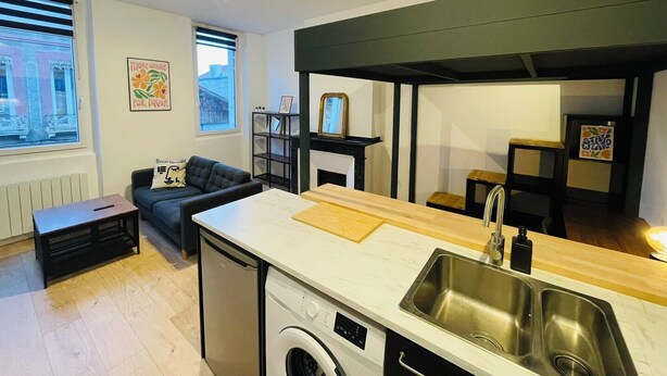 A compact kitchen with a washing machine, a dining area with a sofa, and a view of the city through large windows.