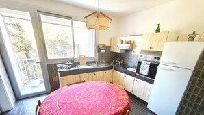 A kitchen with a round table covered in a red tablecloth, wooden cabinets, a white refrigerator, and a window overlooking a balcony. - Beautiful T4 apt with a view of the Gulf d'Ajaccio (Ajaccio)
