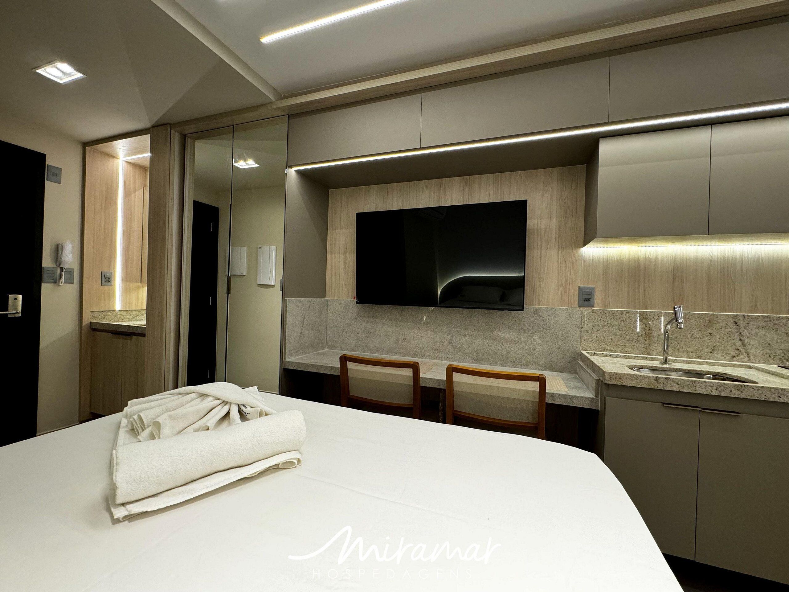 Habitación de hotel con cama, televisor montado en la pared, escritorio con sillas, lavamanos y espejo.