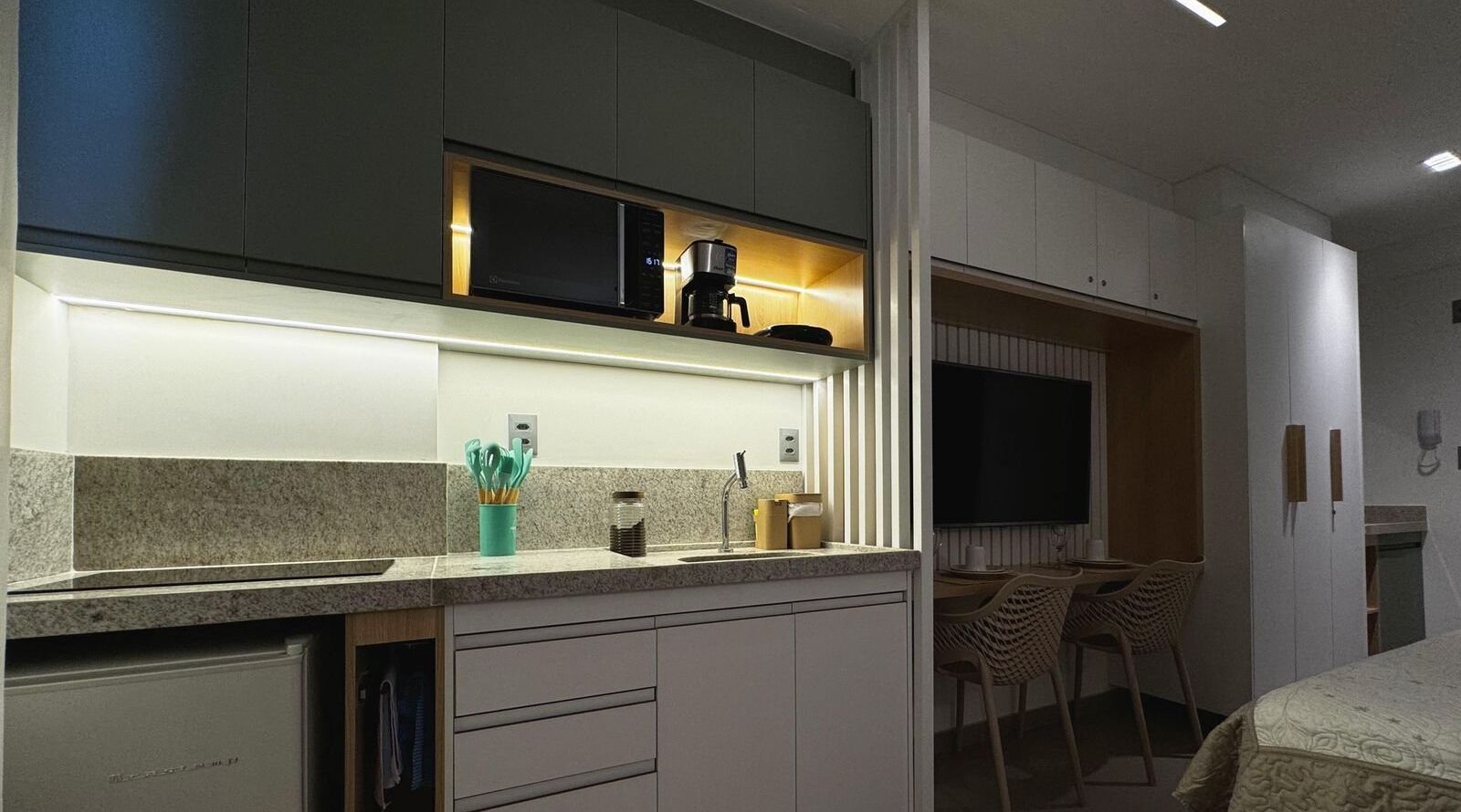 A modern kitchen with a built-in microwave, coffee maker, and a small sink. There are cabinets, a countertop with kitchen utensils, and a dining area with chairs.