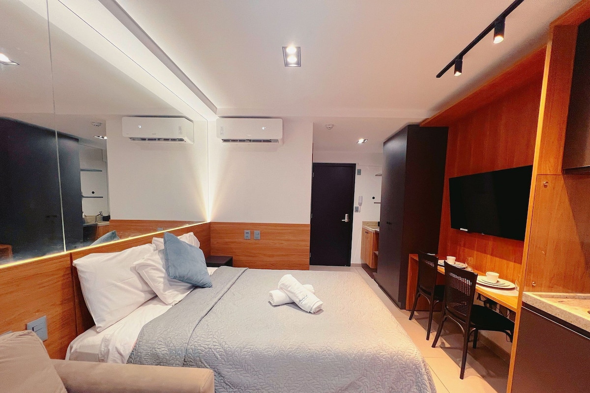 A hotel room with a bed, bedside table, a desk with chairs, a flat-screen TV, and an air conditioning unit.