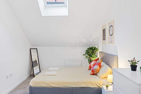A bedroom with a skylight, a bed with pillows, a bedside table with a lamp, and a plant.