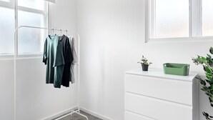 A room with a white wall, a window, a drying rack with clothes, a white dresser, and a green plant.