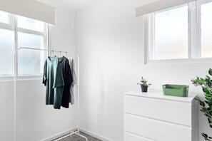 Een kamer met een witte muur, een raam, een droogrek met kleding, een witte dressoir en een groene plant.