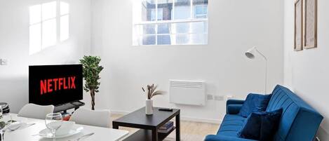 Ein modernes Wohnzimmer mit Flachbildfernseher, einem Esstisch, einem blauen Sofa und einem Fenster mit Jalousien.