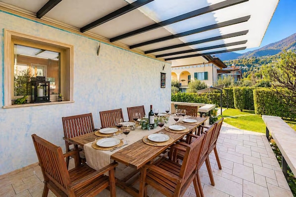 Un espace de restauration en plein air avec une table dressée pour six personnes, entourée de chaises en bois, sous une pergola offrant une vue sur un paysage montagneux.