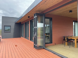 Una casa moderna con terraza de madera, puertas correderas de vidrio grandes y un comedor con mesa y sillas.