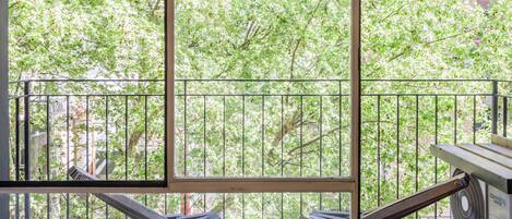A balcony with two wooden benches and a table, overlooking a tree-lined street.
