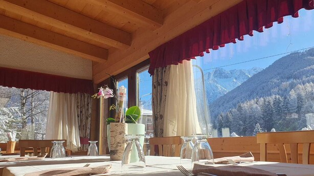 A dining area with a table set for two, wooden beams on the ceiling, and a mountain view through large windows. - Al Maniero (Ponte di Legno)