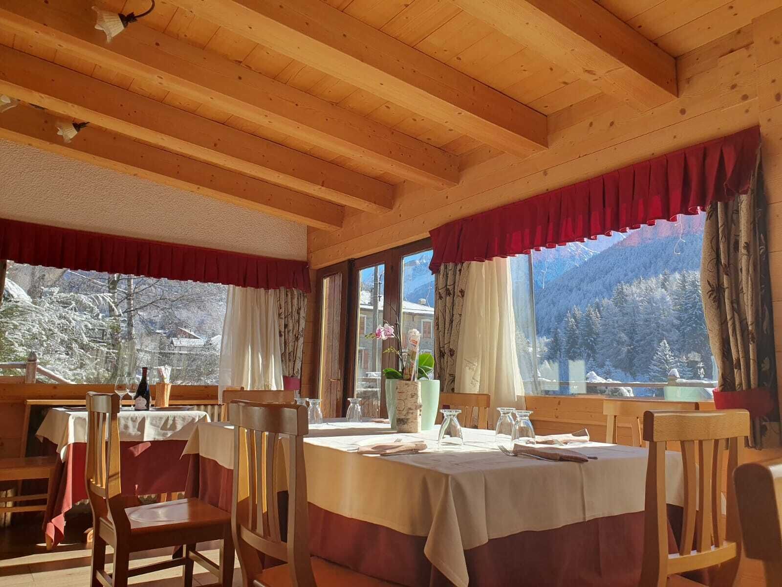 Een houten interieur met gedekte tafels, grote ramen met gordijnen en uitzicht op besneeuwde bergen.