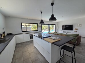 Une cuisine moderne dotée d’un îlot central, d’un coin repas avec des tabourets de bar, d’un téléviseur fixé au mur et d’une grande fenêtre donnant sur un jardin.
